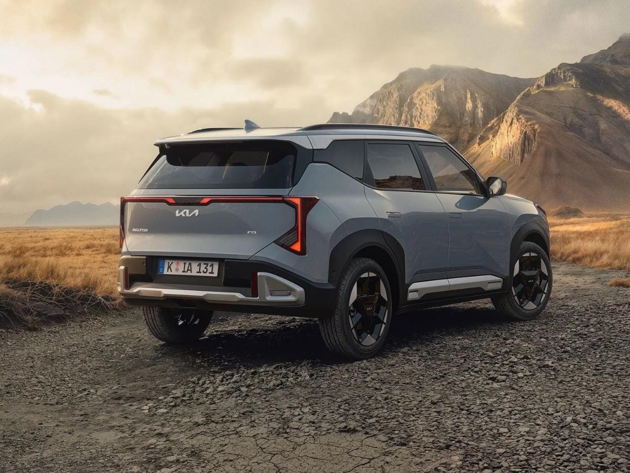Kia Seltos SUV in Grau steht auf einem steinigen Untergrund in einer bergigen Landschaft bei Sonnenuntergang. Das Fahrzeug ist von hinten seitlich zu sehen, mit markanten LED-Rückleuchten und robustem Design vor natürlicher Kulisse.